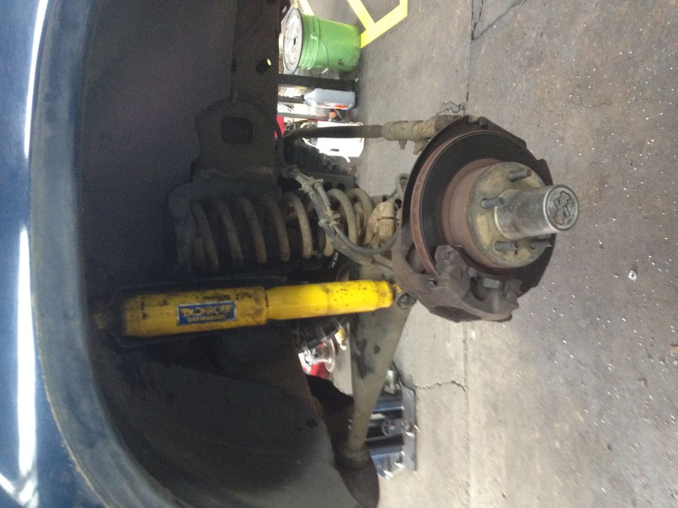 Front suspension with wheel off: brake rotor, hub, coil spring, and yellow Monroe shock in a garage.