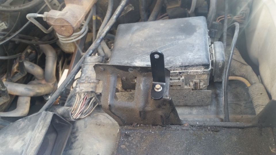 Custom black metal bracket attached to a vehicle fuse box in engine bay.