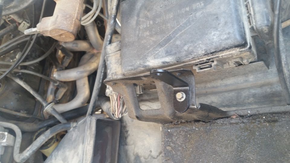 Black metal bracket attached to a black power distribution box in a vehicle engine bay.