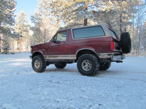 1996 Ford Bronco - A maroon Ford Bronco with a tan top is parked in a snowy forest clearing. The vehicle has large off-road tires and a spare tire mounted on…