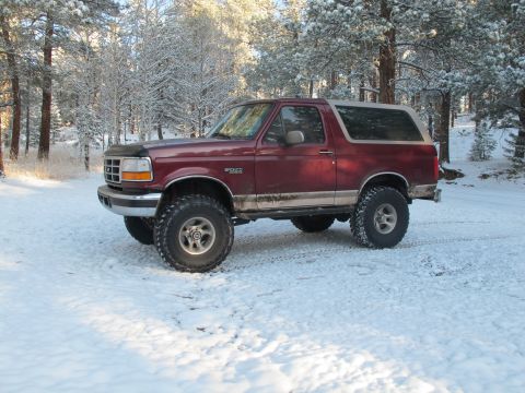 1996 Ford Bronco - A lifted burgundy 1996 Ford Bronco is parked on a snow-covered ground with pine trees in the background. The vehicle has large off-road tir…