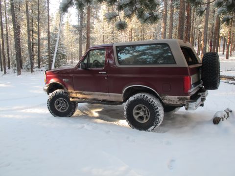 1996 Ford Bronco - A maroon 1996 Ford Bronco with a tan roof sits parked in a snowy forest. The vehicle has large off-road tires and a spare tire mounted on t…