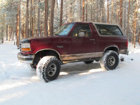 1996 Ford Bronco - A maroon Ford Bronco with large off-road tires sits parked in a snowy forest with tall pine trees. The vehicle has a tan removable top and…