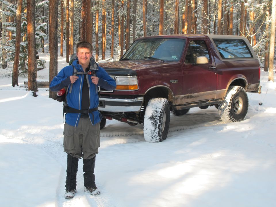 Maroon Ford Bronco with large tires parked in a snowy forest with a person standing in front.