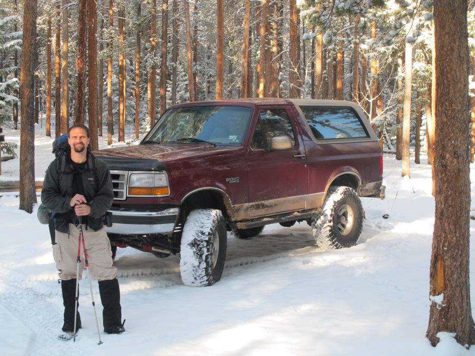 Man standing next to a maroon Ford Bronco in a snowy forest.