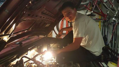 1996 Ford Bronco - A person works on a car's engine bay, with bicycles and other items visible in the background.