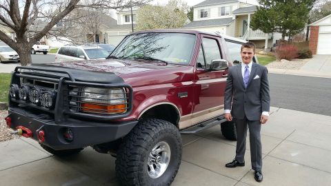 1996 Ford Bronco - A young man in a suit stands beside a lifted red Ford Bronco with a custom bumper and lights.