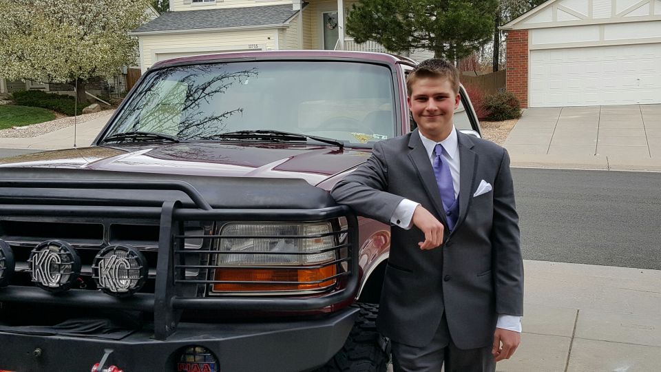 Maroon Ford Bronco with a black bull bar and KC lights, parked in a driveway with a young man in a suit