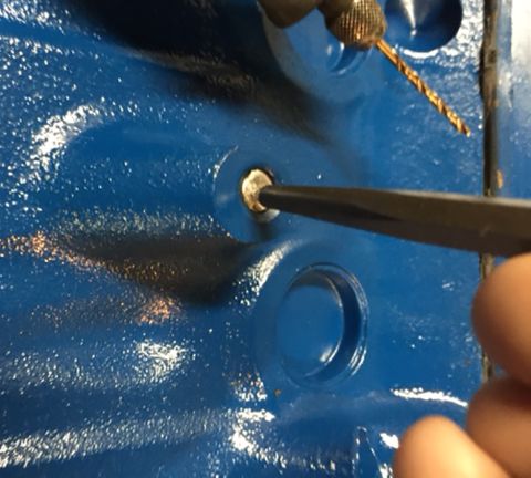 1978 Ford Bronco - A close-up shot shows a drill bit and a screwdriver being used on a blue metal surface, likely a vehicle engine block, to work on a small b…