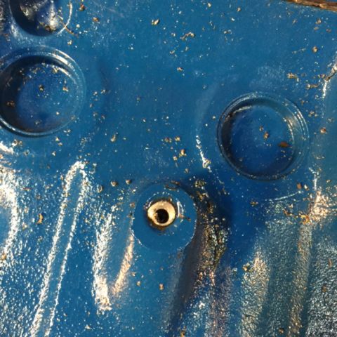 1978 Ford Bronco - A close-up view of a blue painted metal surface with several circular indentations and a central threaded hole. Small flecks of gold debris…