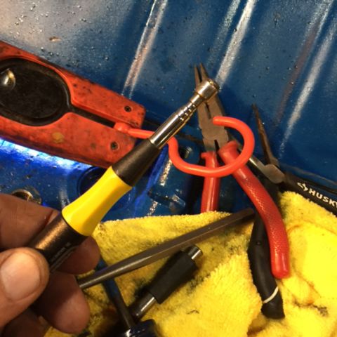 1978 Ford Bronco - A hand holds a magnetic screwdriver with a yellow and black handle. Several tools, including pliers and a red handled tool, are resting on…