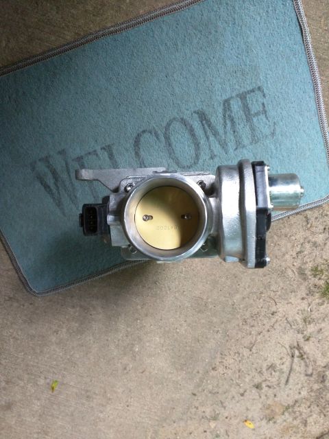 2007 Ford Crown Victoria - A silver throttle body is shown from a top-down perspective, resting on a blue welcome mat placed on a concrete surface. The throttle body…
