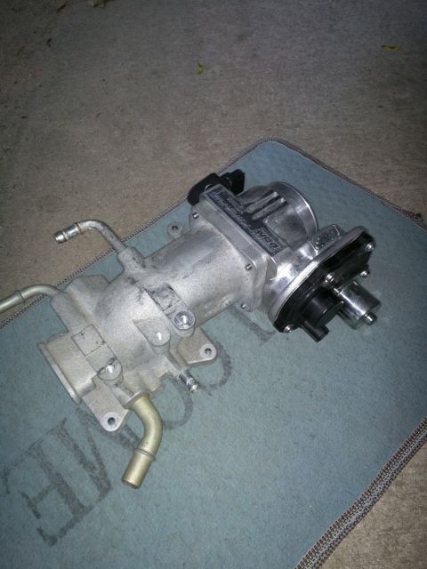 2007 Ford Crown Victoria - A silver throttle body assembly with black connectors and tubing is shown resting on a blue mat on a concrete surface. The assembly feature…