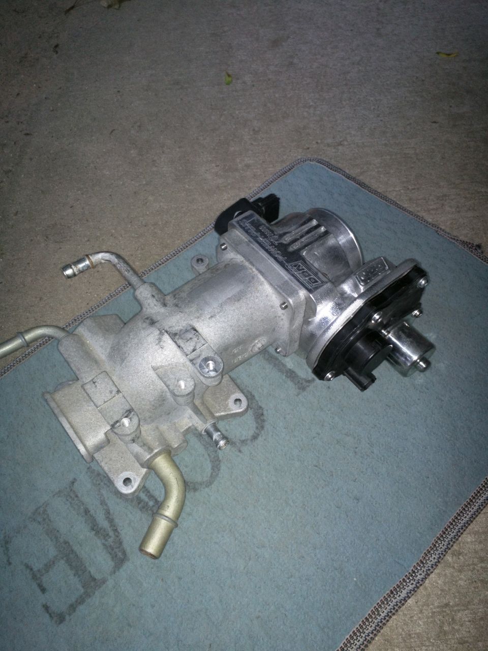 Silver BBK Power Plus throttle body assembly with black connectors and tubing on a blue mat.