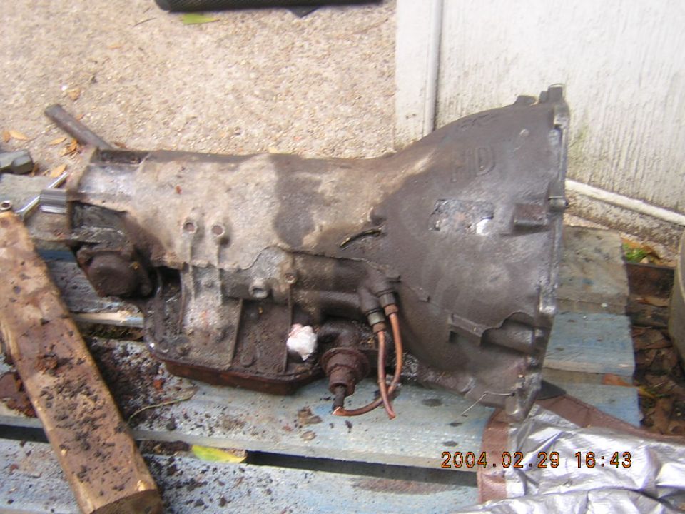 Dirty automatic transmission part with copper lines and a sensor, resting on a wooden pallet