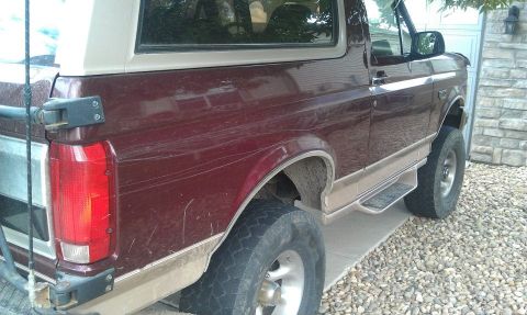 1996 Ford Bronco - The rear driver's side of a dark red Ford Bronco is shown parked on a gravel driveway next to a stone-clad garage. Scratches are visible on…