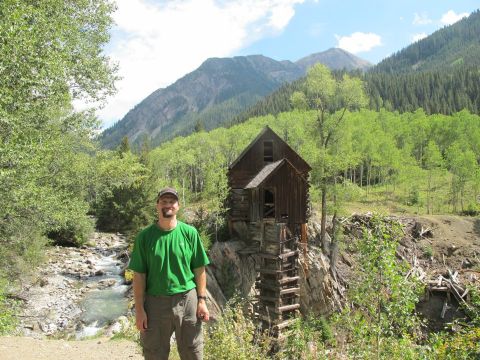 1996 Ford Bronco - A man stands in front of an old wooden structure built on a rocky cliff face, with a stream flowing to his left and mountains covered in tr…