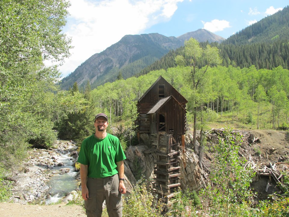 Man standing in front of an old wooden building on a cliff with a stream and mountains.