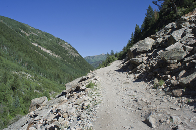 1996 Ford Bronco - A dirt path winds through a rocky mountain landscape with a steep, tree-covered slope on the left and a rocky mountainside on the right.