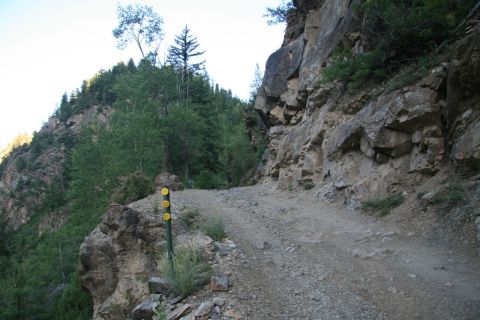 1996 Ford Bronco - A narrow, rocky trail winds along a steep cliff face, with dense green trees on the opposite mountainside. A trail marker with three yellow…