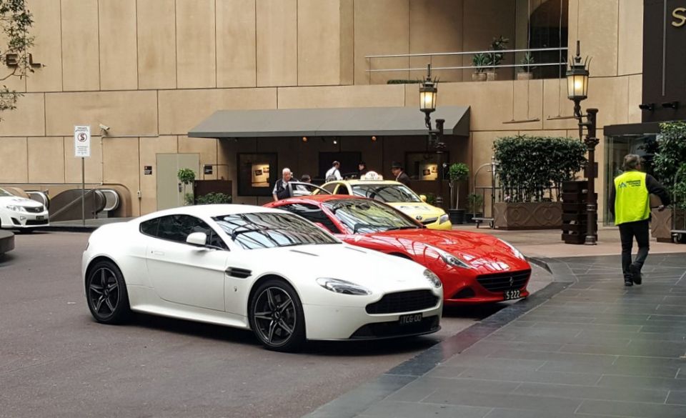 White sports coupe parked in front of a hotel with a red sports coupe and yellow taxi behind it.