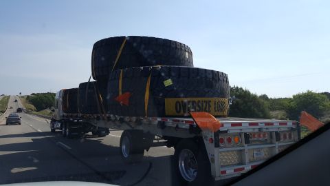 1996 Ford Bronco - A flatbed trailer carries three oversized tires on a highway. The tires are secured with yellow straps and have an 'OVERSIZE LOAD' banner o…