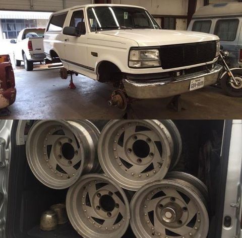1996 Ford Bronco - A white Ford Bronco is on jack stands with its front wheels removed, revealing the brake rotors. Below, a collection of aftermarket aluminu…