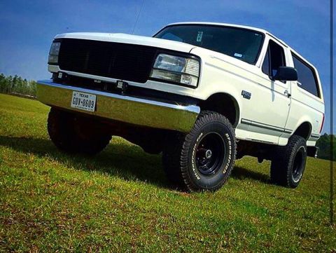 1996 Ford Bronco - A lifted white 1996 Ford Bronco is parked on a grassy hill under a clear blue sky. The front of the vehicle is angled towards the camera, s…