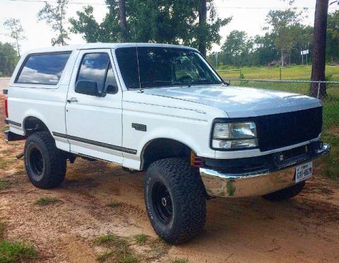 1996 Ford Bronco - A white 1996 Ford Bronco is parked on a dirt road with trees in the background. The vehicle has a chrome bumper and black grille.