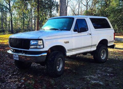 1996 Ford Bronco - A white 1990s Ford Bronco is parked on a dirt lot with trees in the background. The SUV has a black grille, chrome bumper, and large off-ro…