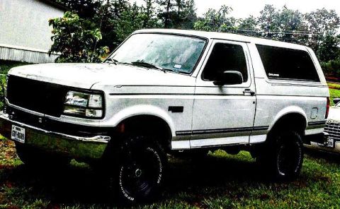1996 Ford Bronco - A white 1990s Ford Bronco is parked on a grassy area with trees in the background. The front of the SUV is visible, showing its grille, hea…