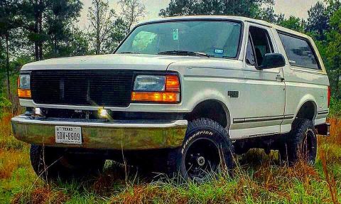 1996 Ford Bronco - A white 1990s Ford Bronco is parked in a grassy field with trees in the background. The front of the SUV is angled towards the camera, show…