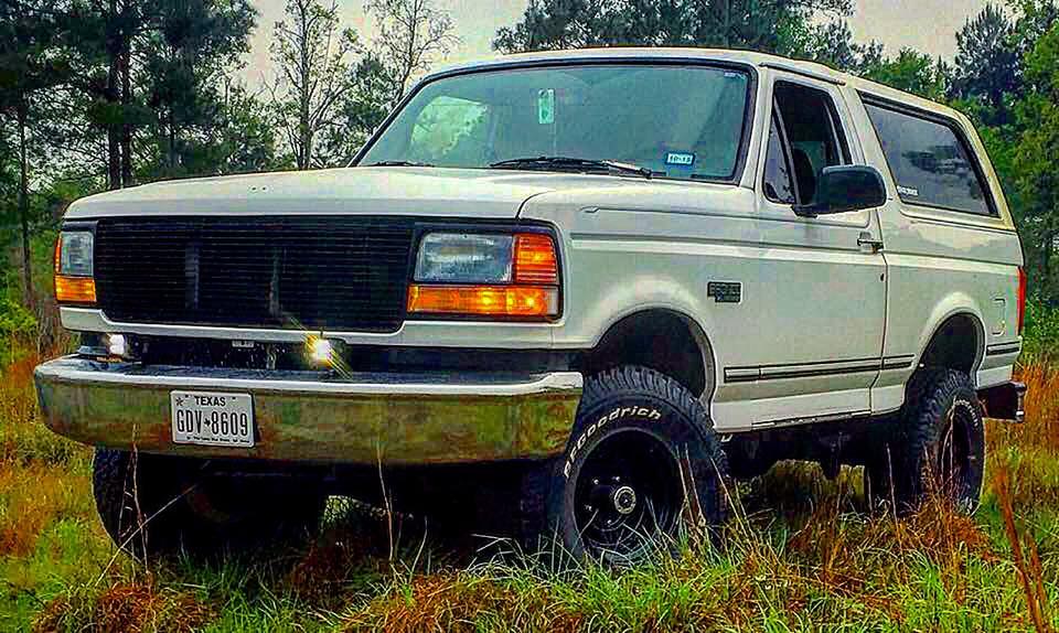 White 1990s Ford Bronco parked in a grassy field, front 3/4 view.