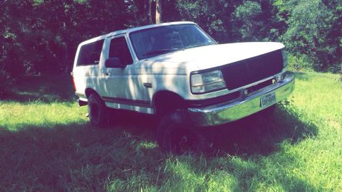 1996 Ford Bronco - A white 1990s Ford Bronco is parked on a grassy area with trees in the background. The front of the SUV is angled towards the camera, showi…