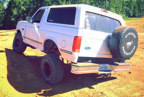 1996 Ford Bronco - The rear passenger side of a lifted white Ford Bronco is shown parked on a dirt lot with trees in the background. It features a spare tire…