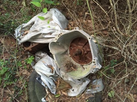 1996 Ford Bronco - A rusty automatic transmission bell housing and torque converter are partially covered by a silver tarp, lying on the ground amidst brush a…