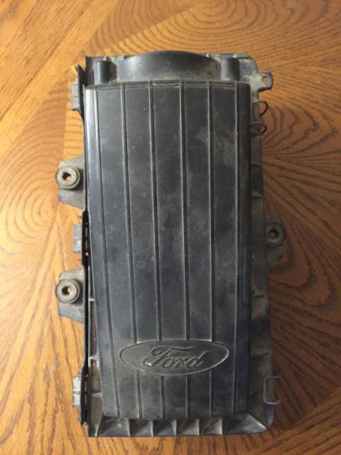 1996 Ford Bronco - A black Ford air cleaner housing is shown from a top-down perspective, resting on a wooden surface. The Ford logo is visible in raised lett…
