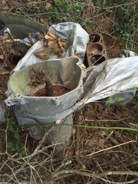 1996 Ford Bronco - A disassembled automatic transmission housing and a rusted engine block are partially covered by a tarp, resting on the ground amidst dry l…