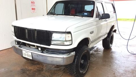1996 Ford Bronco - A white 1990s Ford Bronco is parked on a wet surface, possibly after a wash. The front of the SUV is visible, showing its grille, headlight…