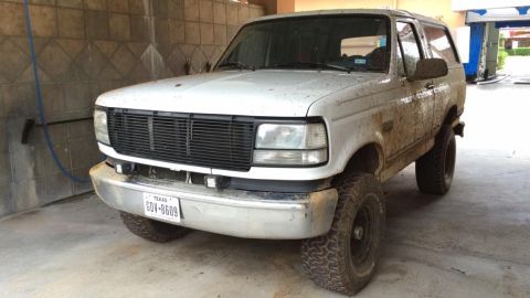 1996 Ford Bronco - A dirty white 1990s Ford Bronco is parked in a car wash bay. The front of the SUV is visible, showing its grille, headlights, and Texas lic…