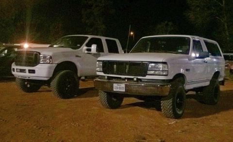 1996 Ford Bronco - Two lifted white Ford trucks are parked on a dirt lot at night. The truck on the right is a 1996 Ford Bronco with a Texas license plate rea…