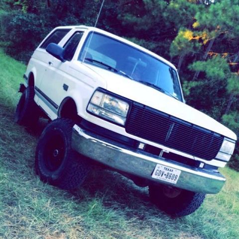 1996 Ford Bronco - A white 1990s Ford Bronco is parked on a grassy slope with trees in the background. The vehicle features large off-road tires and a chrome…