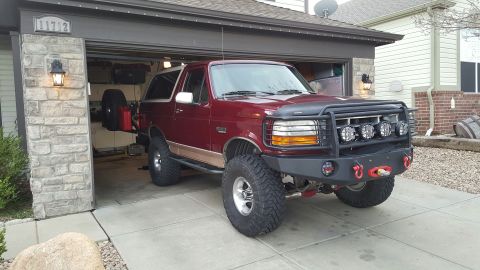 1996 Ford Bronco - A maroon Ford Bronco with a large aftermarket bumper and winch is parked in front of an open garage. The vehicle has large off-road tires a…