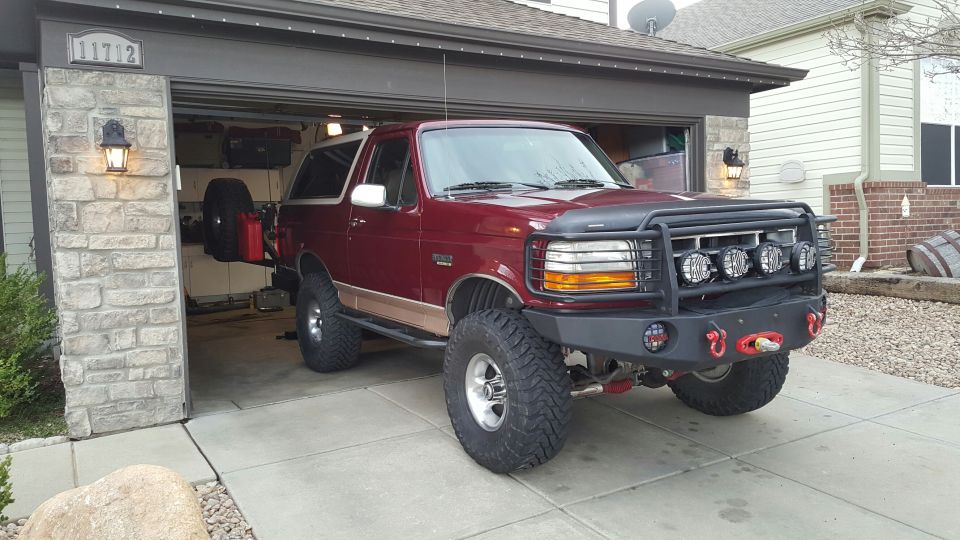 Maroon Ford Bronco with aftermarket bumper and winch parked in front of an open garage.