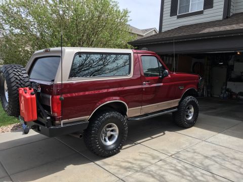 1996 Ford Bronco - The rear passenger side of a red and tan Ford Bronco is parked in a driveway in front of a garage. The vehicle has a spare tire mounted on…