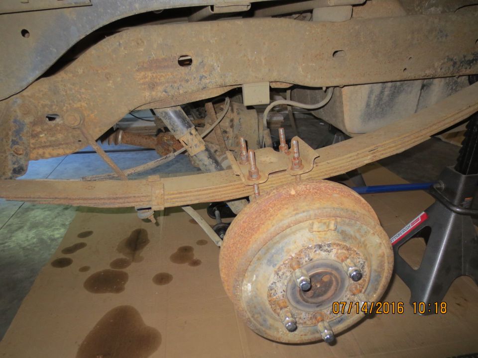 Underside of a vehicle showing rusty leaf springs and brake drum assembly.