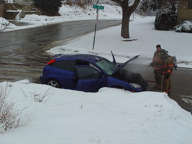 Crashed blue Ford Focus hatchback in snow-filled ditch with two firefighters