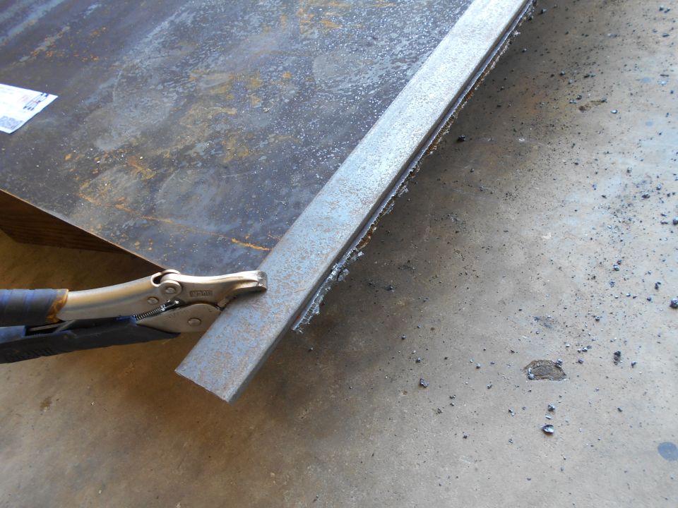 Metal bar held by locking pliers against sheet metal, with shavings on concrete.