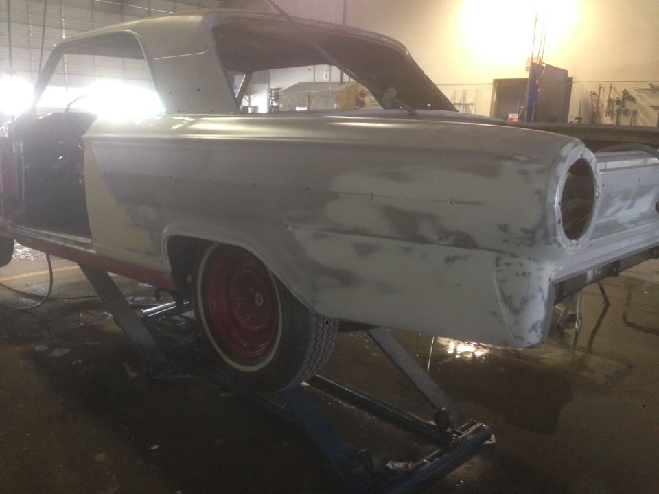 1964 Ford Fairlane in primer on a lift, showing bodywork and a red wheel with whitewall tire