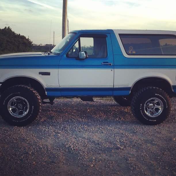 Side view of a blue and white Ford Bronco with aftermarket wheels and tires on a gravel lot.